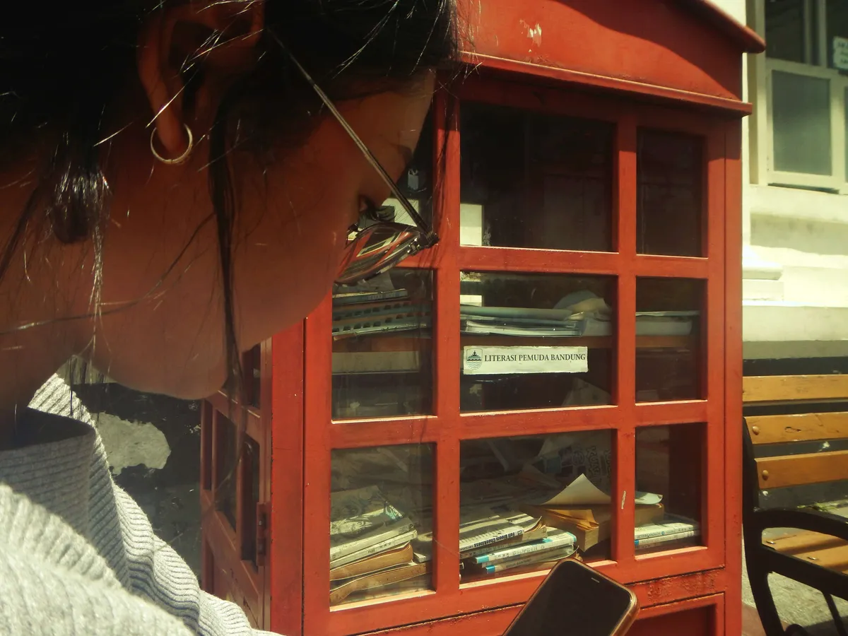 Seorang pengunjung duduk di dekat Street Library sambil menatap gawai, sementara buku-buku di dalam kotak baca dibiarkan menumpuk dan berdebu. Pemandangan ini mencerminkan tantangan literasi Kota Bandung. (Sumber: Adellia Ramadhani)