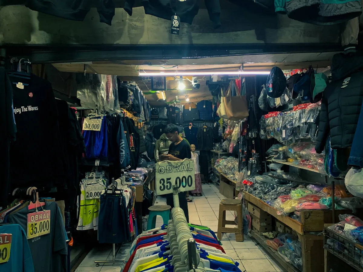 Dimas Angga sedang melayani pembeli di pasar tradisional, dengan berbagai pilihan pakaian dan aksesoris yang dijual dengan harga terjangkau. Foto ini diambil di sebuah toko pakaian di kawasan Bandung, yang terkenal dengan produk-produk lokal berkualitas. (Sumber: Tito Andrean | Foto: Tito Andrean)