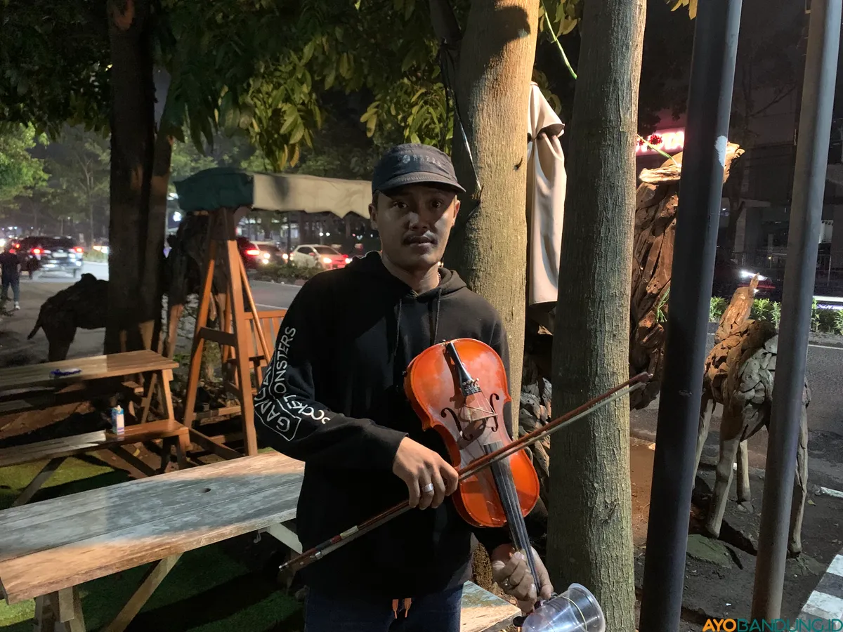 Ricky Rustandi Pratama memainkan biola di persimpangan jalan untuk menambah penghasilan sambil bekerja sebagai cleaning service. (Sumber: ayobandung.id | Foto: Halwa Raudhatul)