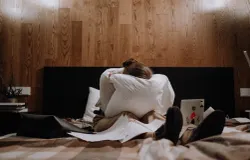 Seorang wanita duduk di atas tempat tidur sambil memegang bantal, dikelilingi kertas dan laptop, menunjukkan ekspresi stress. (Sumber: pexels | Foto: Ron Lach)