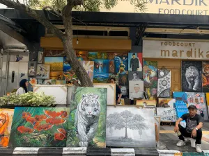 Ian seorang pelukis lokal dan karya lukisannya yang dipajang di trotoar Jalan Braga. (Sumber: ayobandung.id | Foto: Toni Hermawan)