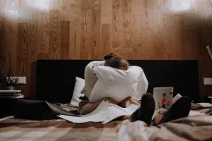 Seorang wanita duduk di atas tempat tidur sambil memegang bantal, dikelilingi kertas dan laptop, menunjukkan ekspresi stress. (Sumber: pexels | Foto: Ron Lach)